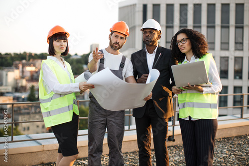 Group of multiethnic industrial employees planning building project and meeting on panoramic terrace. Four international colleagues using computer and technical plan for remodeling business center.