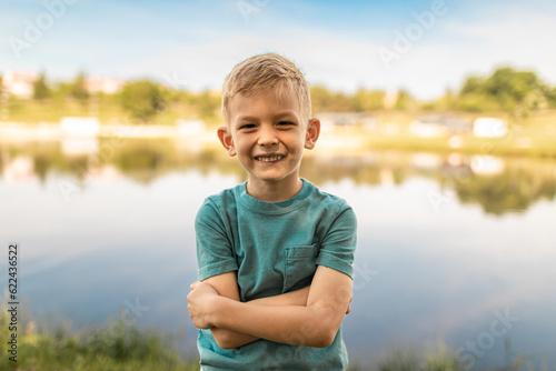 Happy blond boy in casual clothing, smiling at camera by a serene lake.
