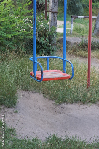 one empty swing seat made of blue metal and red boards on the street against the background of green grass