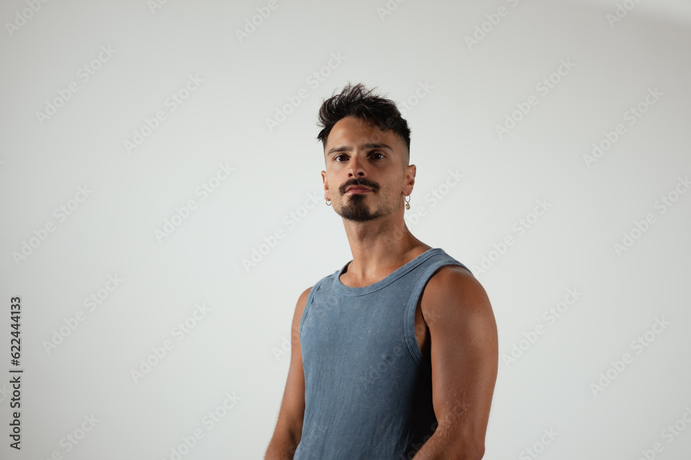 A man stands for a casual pose and looks to the side, gazing directly at the camera. His expression is neutral, with some seriousness. He is wearing a blue tank top, showing his athletic physique. A man stands for a casual pose and looks to the side, gazing directly at the camera. His expression is neutral, with some seriousness. He is wearing a blue tank top, showing his athletic physique.