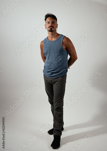 A young man poses for a portrait in a white studio. He is wearing a blue tank top, gray jeans, and black shoes.  © Levi Meir Clancy