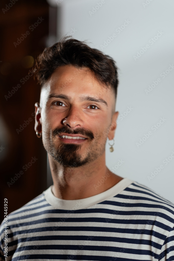 A handsome man smiles for a portrait, wearing a striped shirt and earrings. He is posing in an indoor studio space. A handsome man smiles for a portrait, wearing a striped shirt and earrings. He is posing in an indoor studio space.