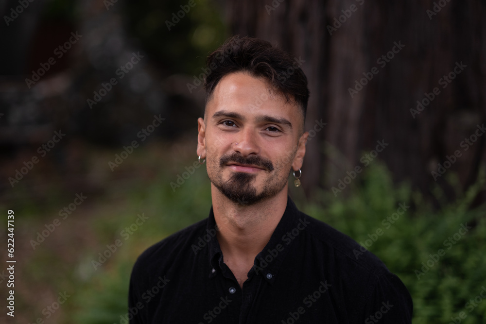 A man smiles while standing in a park. In the background, there is a redwood, some greenery, and a structure in the distance. A man smiles while standing in a park. In the background, there is a redwood, some greenery, and a structure in the distance.