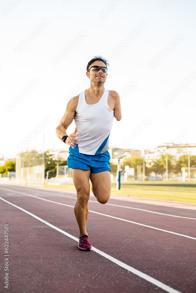 An adult runner with an amputated arm is running down an outdoor running track. The man is wearing a watch and sportswear. Concept of disabled athletes, improvement and motivation in sport.