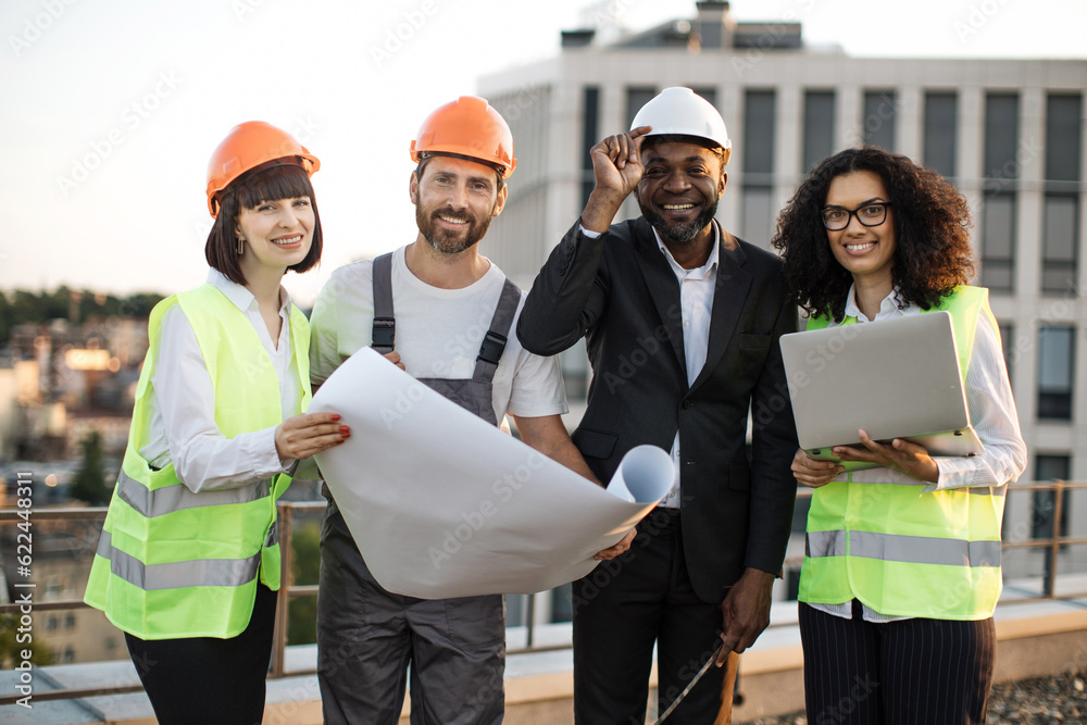 Portrait of multicultural group of developers posing on camera with portable gadget and blueprints. Four international colleagues in vests and hardhats smiling while meeting at panoramic terrace. Portrait of multicultural group of developers posing on camera with portable gadget and blueprints. Four international colleagues in vests and hardhats smiling while meeting at panoramic terrace.