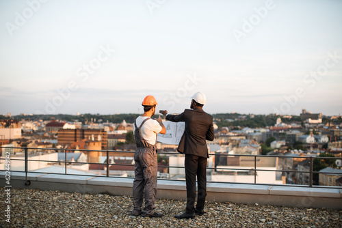 Back view of foreman in grey uniform and business investor in black suit watching together at engineering drawings while standing on panoramic terrace. Male builders discussing real estate project.