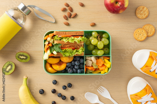 On wooden desk, plastic lunchbox is filled with nutritious elements such as kiwi, apple, banana, peach, grapes, vegetables, sandwich, blueberries, almonds, cookies, water bottle, cutlery, and sneakers