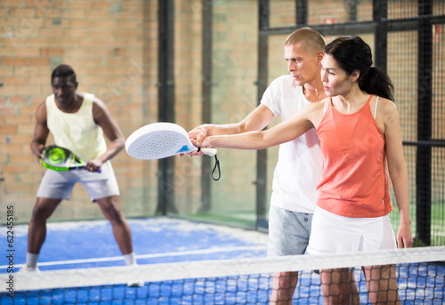 Young man teaching Asian woman to properly hold paddle racquet during training on indoor tennis court © JackF