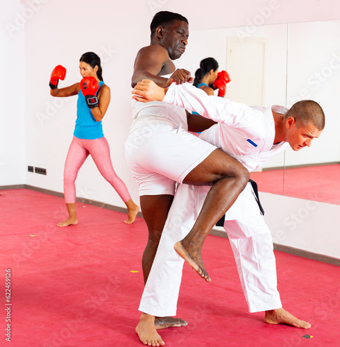 African-american and European men in kimono exercising knee strike in gym. Asian woman boxing behind them.