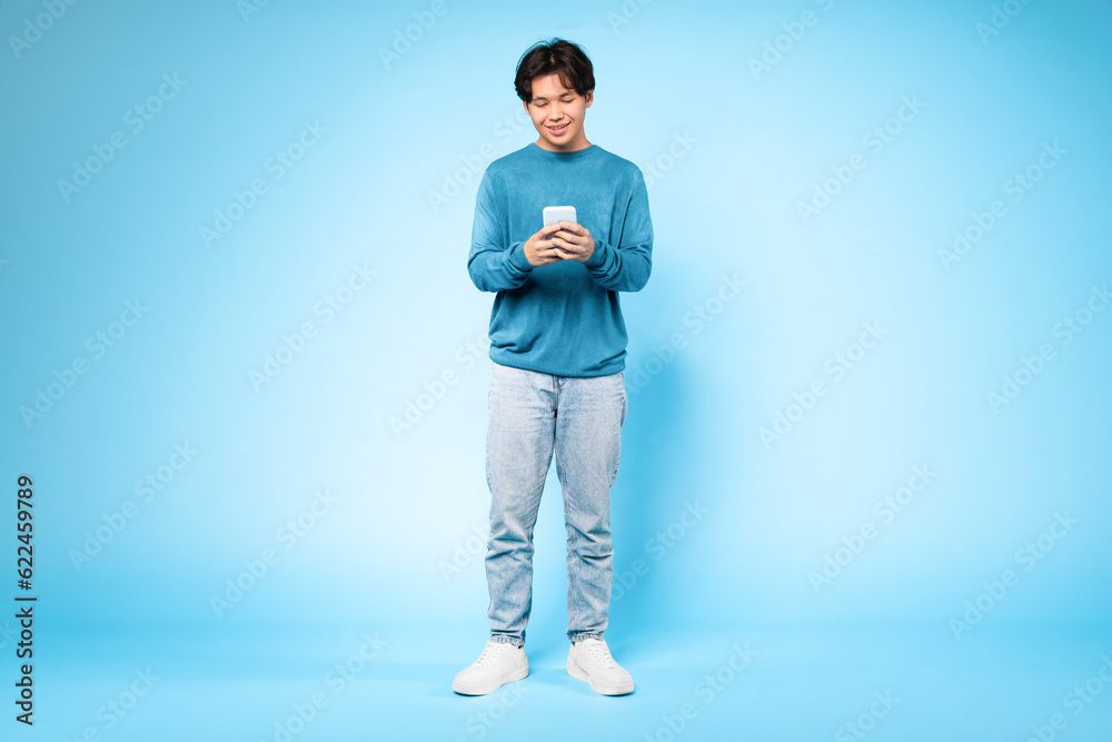 Fototapeta premium Asian guy using smartphone enjoying nice mobile app, blue background