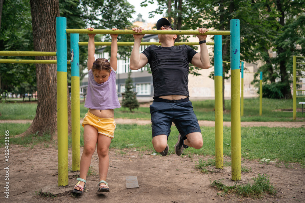 Fototapeta premium Father with child girl doing pull-ups on workout outdoor area. Healthy active lifestyle, happy family time. Modern fatherhood concept 