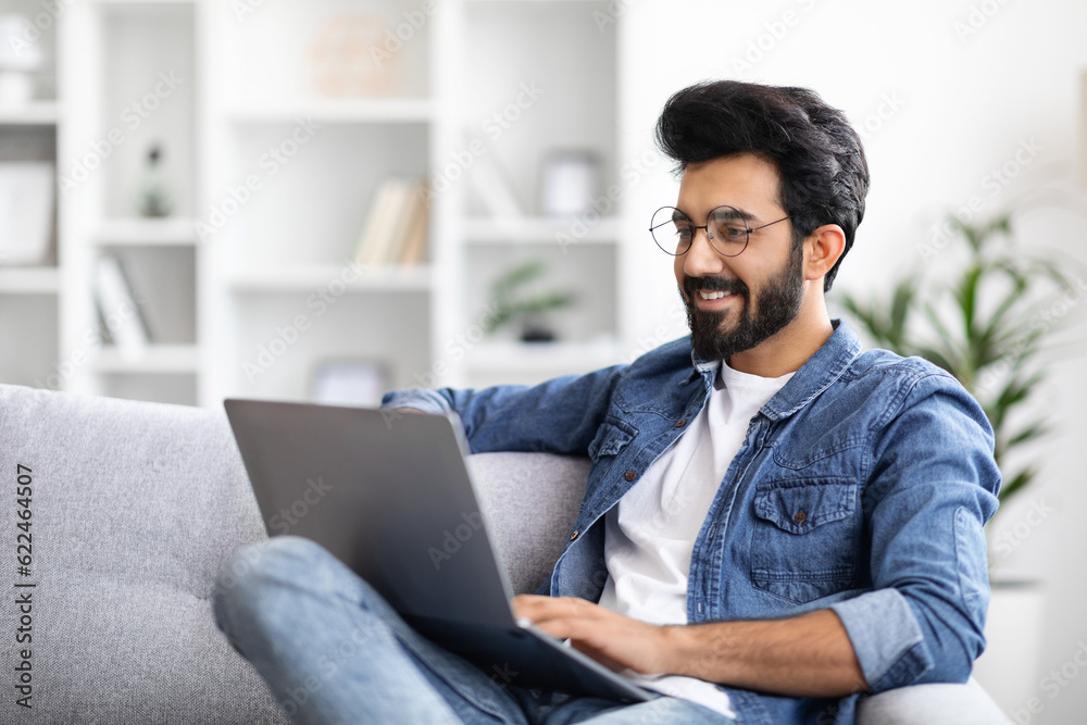 Smiling Indian Man Working On Laptop While Sitting On Couch At Home Smiling Indian Man Working On Laptop While Sitting On Couch At Home