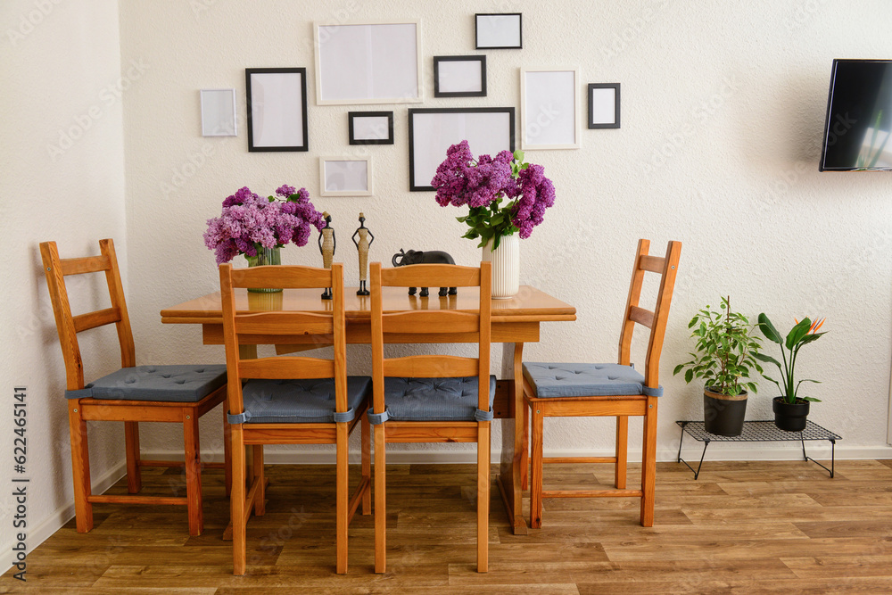 Vases with beautiful lilac flowers on table, chairs and blank pictures in interior of light living room Vases with beautiful lilac flowers on table, chairs and blank pictures in interior of light living room