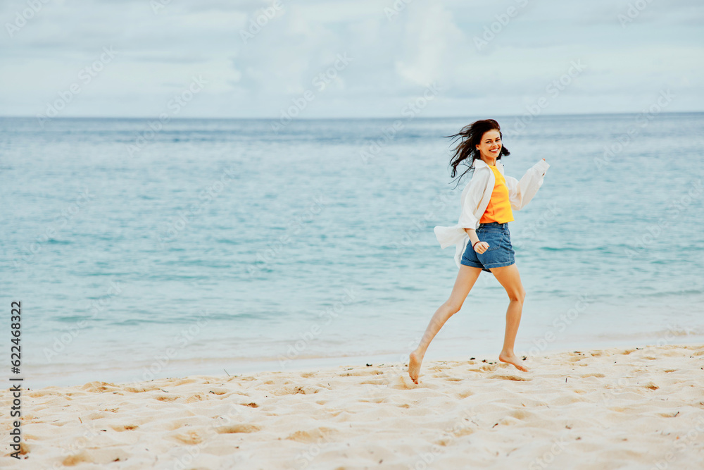 Sports woman runs along the beach in summer clothes on the sand in a yellow T-shirt and denim shorts white shirt flying hair ocean view, beach vacation Sports woman runs along the beach in summer clothes on the sand in a yellow T-shirt and denim shorts white shirt flying hair ocean view, beach vacation