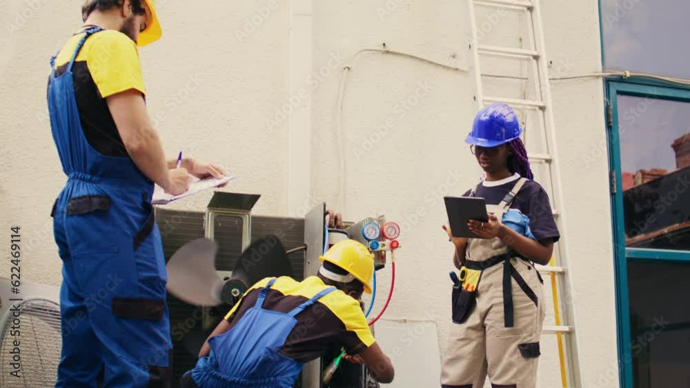 Trained diverse teamworking servicemen cleaning external condenser internal coolants, checking freon level and calibrating thermostats while coworker checks maintenance procedures on tablet Trained diverse teamworking servicemen cleaning external condenser internal coolants, checking freon level and calibrating thermostats while coworker checks maintenance procedures on tablet
