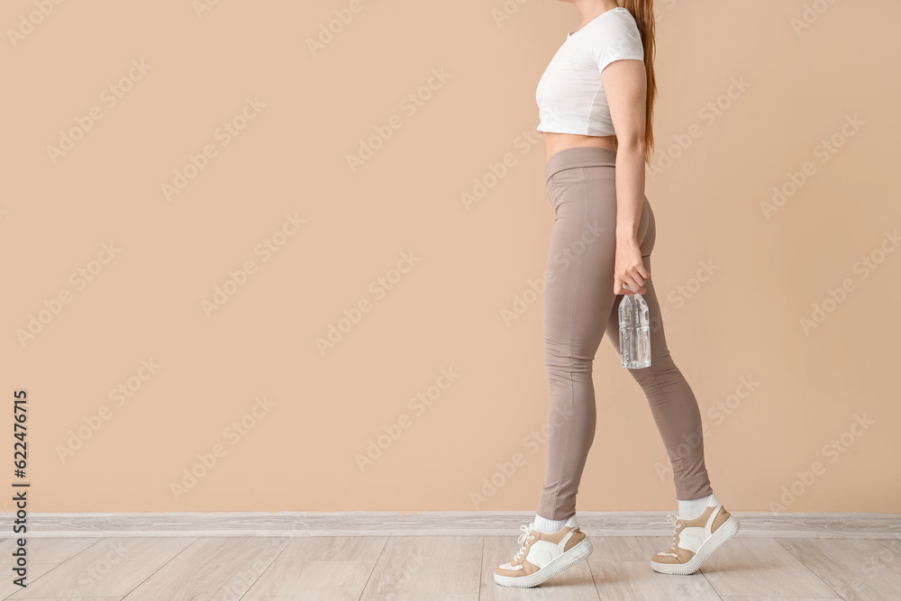 Young woman in sportswear and with bottle of water near beige wall Young woman in sportswear and with bottle of water near beige wall