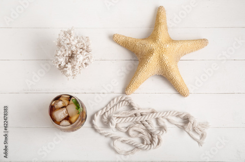 Glass of cold Cuba Libre cocktail, coral and starfish on white wooden background