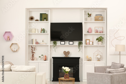 Stylish room interior with beautiful fireplace, TV set, sofa, armchairs and shelves with decor