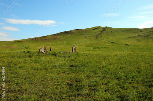 Ancient stone menhirs in a wide clearing overgrown with tall grass at the foot of a high mountain with parallel stone formations.