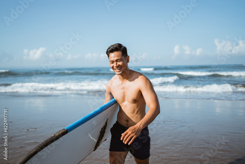 Canvas Print Asian male surfer happily carrying a surfboard after training during a beach wal