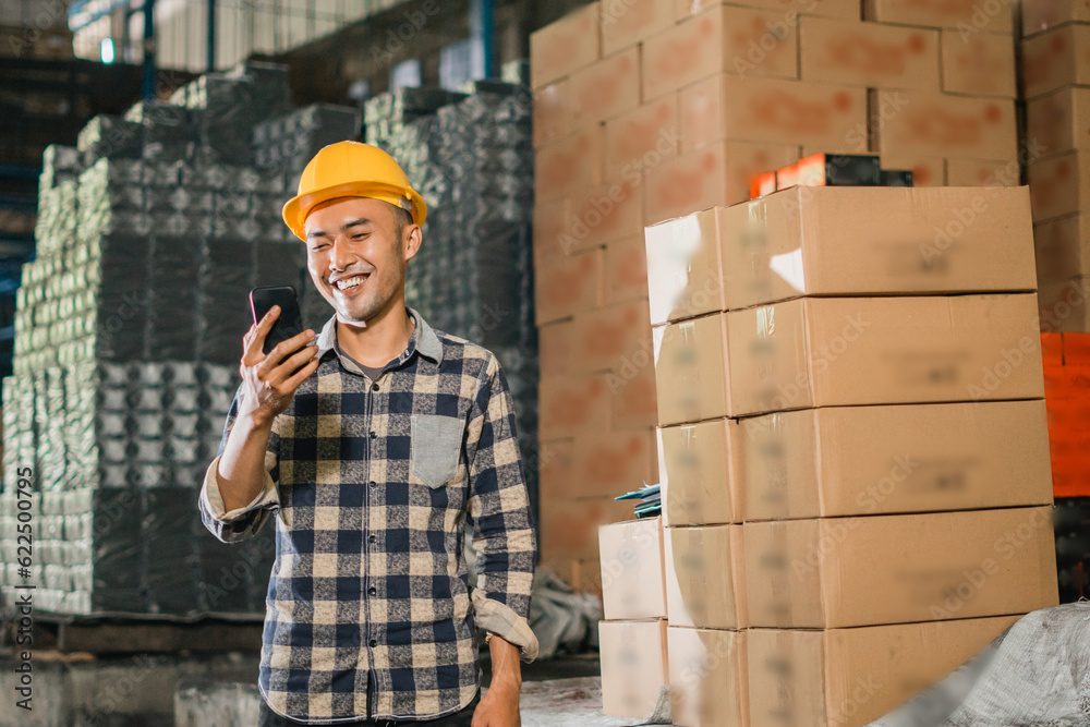 male factory worker wearing safety helmet using cell phone while working in warehouse male factory worker wearing safety helmet using cell phone while working in warehouse