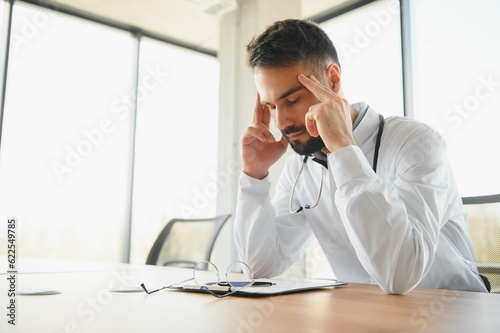 Handsome doctor man wearing medical uniform sitting on his workplace tired holding his head feeling fatigue and headache. Stress and frustration concept