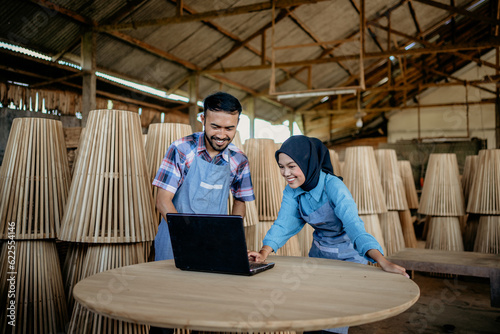 two smiling Asian entrepreneurs using laptop computer together on wooden table at a woodcraft shop