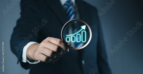 Business investment earning income concept. Businessman hand holding magnifier glass with virtual bar graph with increasing arrow for analysis technical graph of stock market chart.
