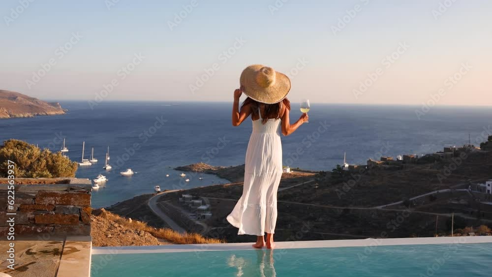 Attractive holiday woman in a white dress at the swimming pool enjoys the sunset over the mediterranean sea with a glass of wine