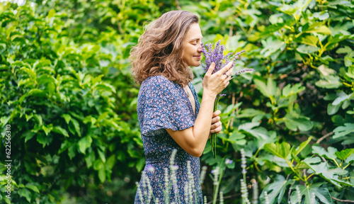 Young happy woman in a dress standing in the garden with a bouquet of lavender and inhaling the scent of flowers