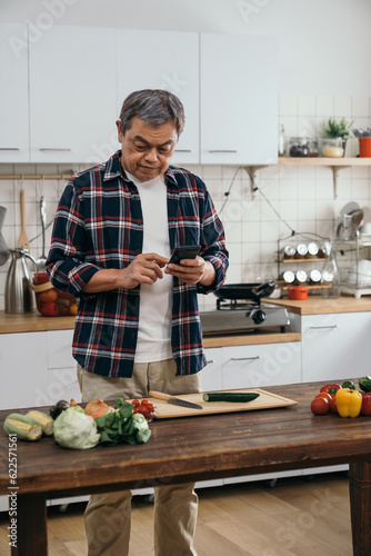 A senior man of Asian descent standing in his kitchen, checking the ingredients on the counter and using his smartphone to find cooking instructions or recipes.