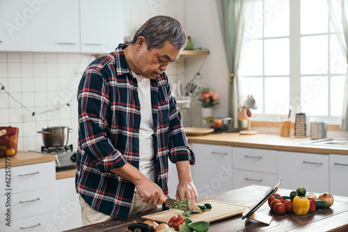 The standing senior Asian man learning to cook with a digital tablet pad, chopping vegetables on the board with ingredients in the home kitchen.