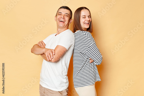 Happy cheerful young couple wife and husband wearing casual attires isolated over beige background standing back to back looking at cmaera and laughing having fun together. photo