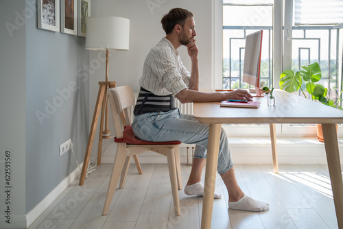 Focused male freelancer wearing lumbar brace sitting at table using computer, young man use orthopedic corset belt while working remotely with herniated disc. Work from home and back pain  photo