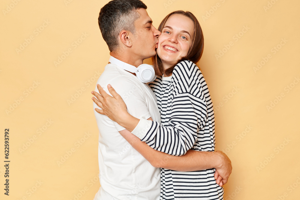 Happy cheerful young couple wife and husband wearing casual attires standing isolated over beige background man kissing woman cheek with love family expressing happiness. Happy cheerful young couple wife and husband wearing casual attires standing isolated over beige background man kissing woman cheek with love family expressing happiness.