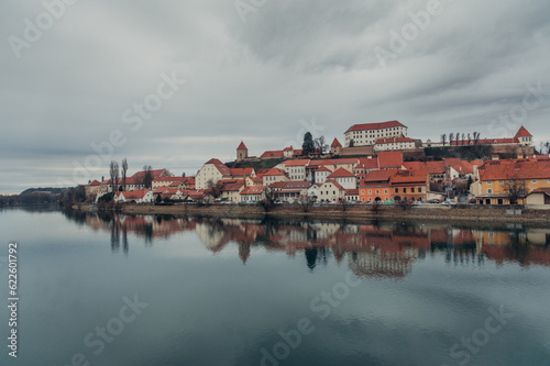 A panoramic view of Ptuj, Slovenia, reveals its charming old town, majestic castle, and a meandering river under a cloudy sky.