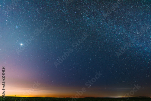 Night Starry Sky With Glowing Stars Above Countryside Field Landscape In Early Spring. Bright Glow Of Planet Venus In Sky Among The Stars. Sky In Warm Lights Of Evening Sunset Dawn Or Morning Sunrise. photo
