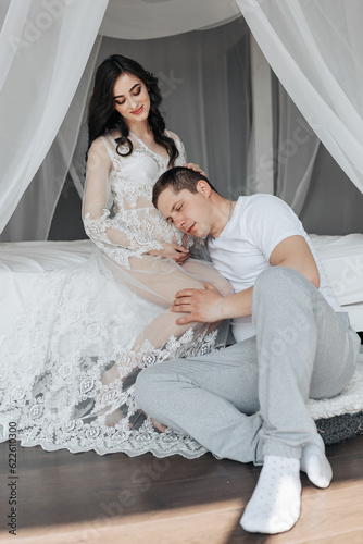 Young pregnant woman on bed in white lace dress holds her pregnant belly, her husband leans his ear against her tummy. Motherhood. Concept of pregnancy, motherhood, preparation and waiting.