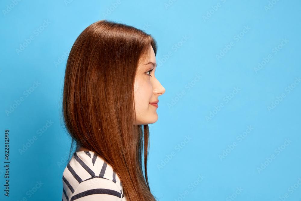Side view portrait of young girl with straight hair looking forward, posing against blue studio background. Concept of human emotions, fashion, youth, lifestyle, female beauty, ad Side view portrait of young girl with straight hair looking forward, posing against blue studio background. Concept of human emotions, fashion, youth, lifestyle, female beauty, ad