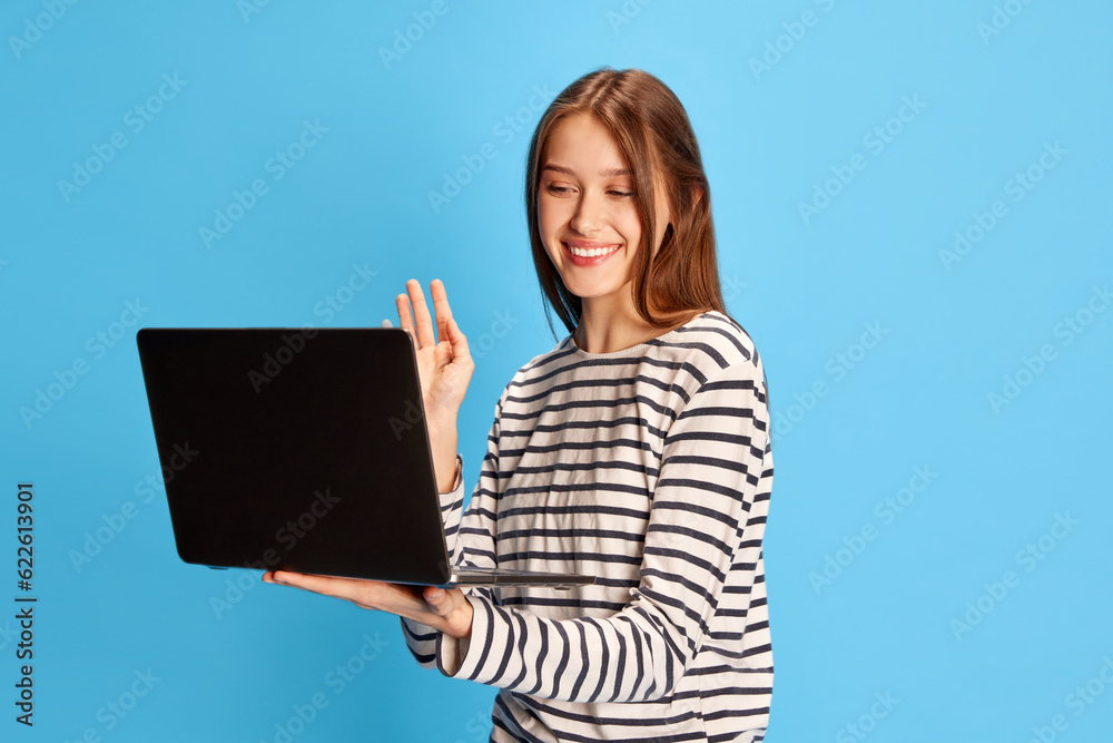 Portrait of young smiling girl, student having online video call via laptop over blue studio background. Communication. Concept of business, education, emotions, fashion, youth, lifestyle, beauty, ad Portrait of young smiling girl, student having online video call via laptop over blue studio background. Communication. Concept of business, education, emotions, fashion, youth, lifestyle, beauty, ad