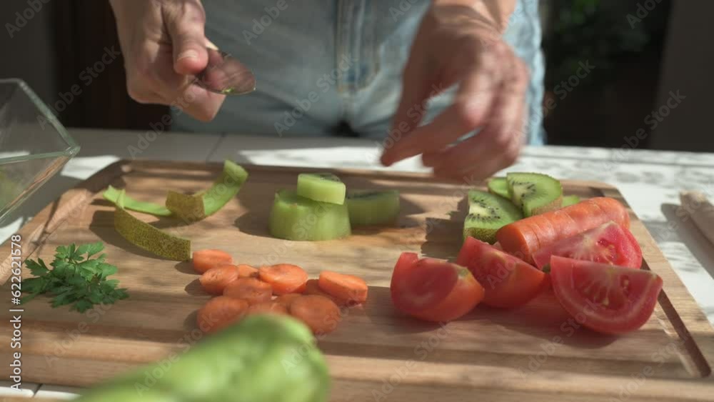 Real time handheld shot of crop anonymous woman peeling juicy kiwi while cutting vegetables and fruits making healthy snacks in kitchen illuminated by sunlight Real time handheld shot of crop anonymous woman peeling juicy kiwi while cutting vegetables and fruits making healthy snacks in kitchen illuminated by sunlight