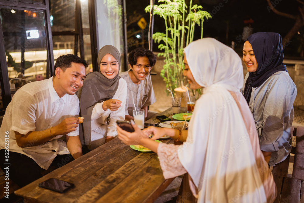 portrait of a group of Muslim friends looking at mobile phones together while breaking fast at an outdoor cafe