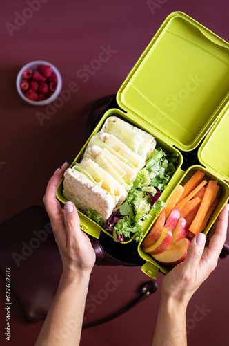 Female hand holding lunch box with sandwiches, carrot sticks, apple, lettuce and raspberries on a burgundy background with a woman's handbag and wallet. Healthy food to go concept. Top view