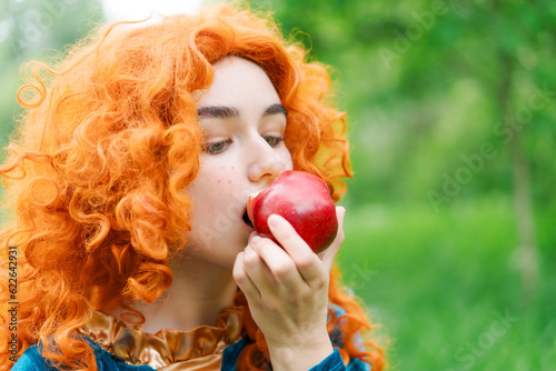 portrait of a young women in a red curly wig eats a big red apple in the park close-up