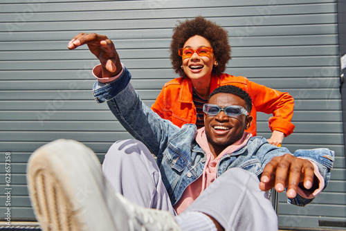 Positive african american woman in sunglasses and bright clothes having fun with stylish best friend in denim jacket sitting in shopping cart on urban street, friends with stylish vibe
