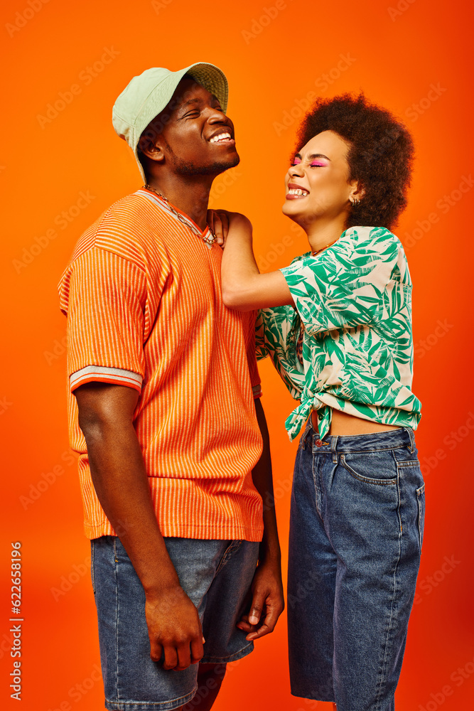 Positive young african american woman with bold makeup and summer outfit hugging stylish best friend in panama hat while standing together isolated on orange, friends in trendy clothes Positive young african american woman with bold makeup and summer outfit hugging stylish best friend in panama hat while standing together isolated on orange, friends in trendy clothes