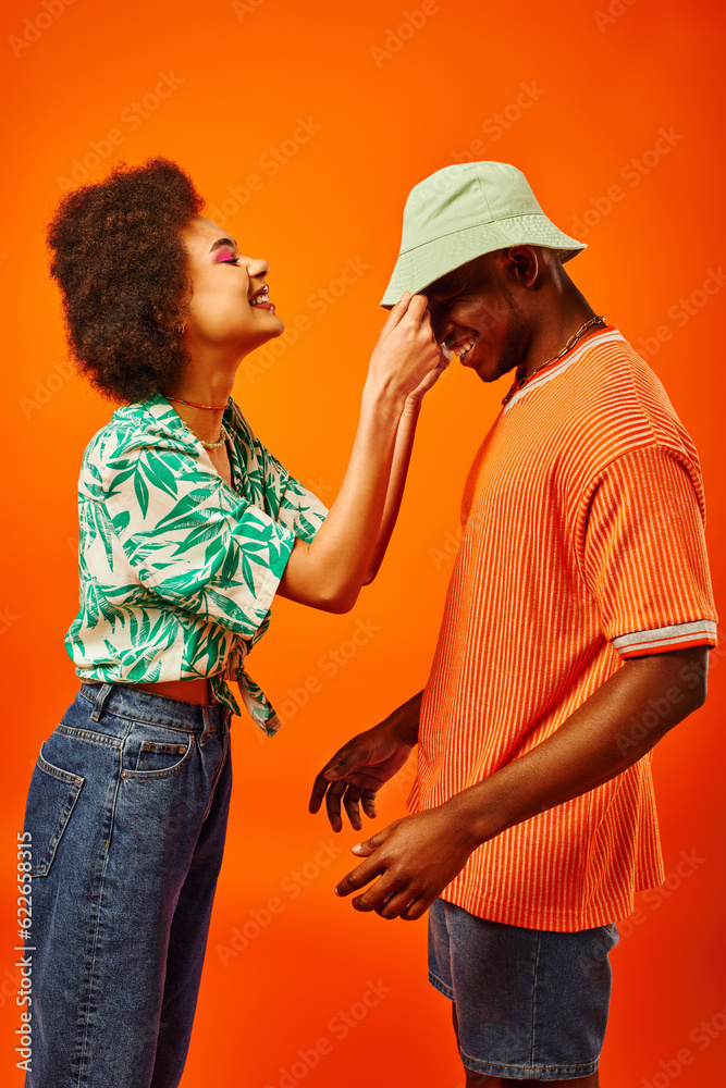 Side view of smiling young african american woman in summer outfit touching panama hat on best friend and having fun together while standing isolated on orange, friends in trendy clothes