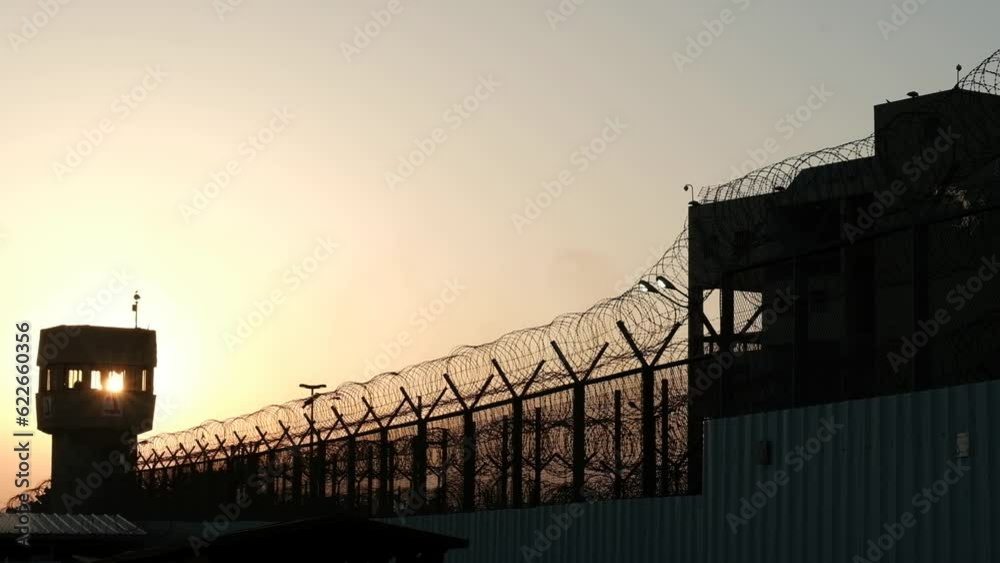 A silhouette of a prison watchtower at sunset. the barbed wire is apparent at the bottom of the frame and the sun ball itself is visible directly through the tower windows.