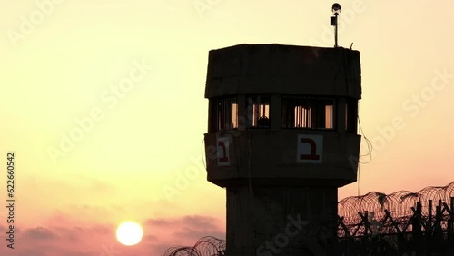 A silhouette of a prison watchtower at sunset. while the sun ball is nicely set at the bottom of the frame, a military helicopter is passing right underneath it. a 4K video clip, Israel.