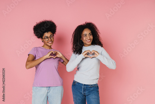 Two excited african women making heart sign with hands while standing isolated over pink background